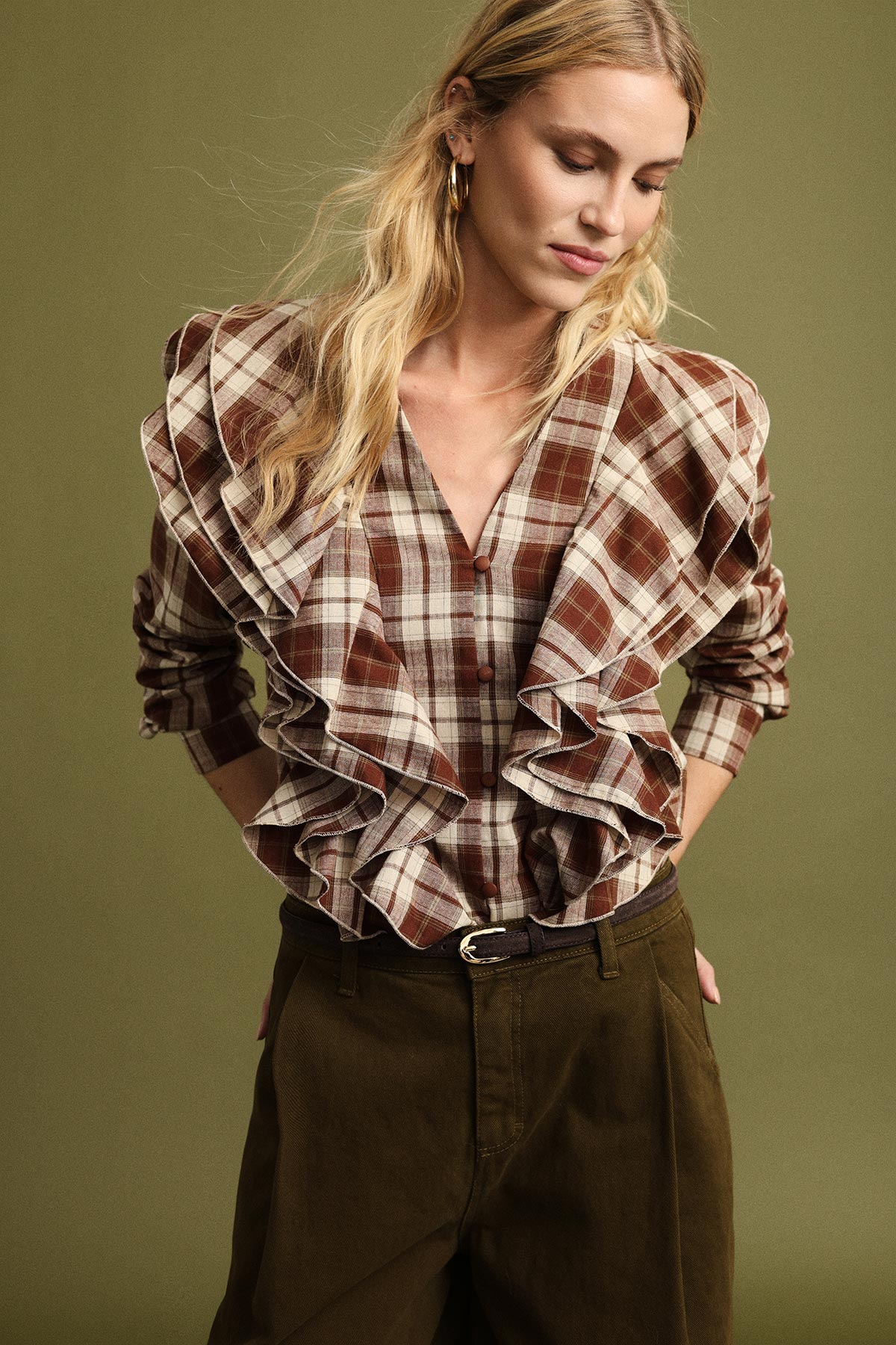 Model wearing brown checked ruffled blouse and olive-coloured high-waist trousers on a muted green background