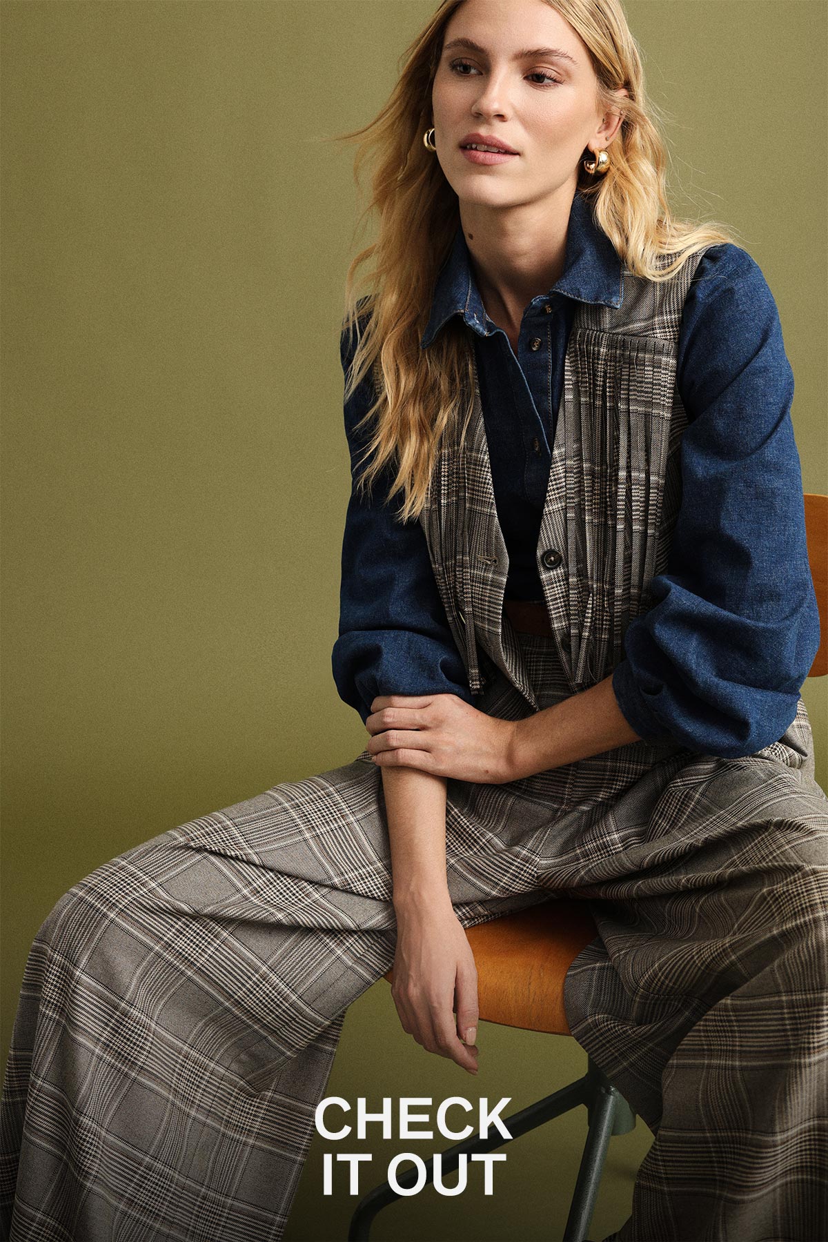 Seated female model wearing denim shirt, checked waistcoat and wide-leg checked trousers on green background with 'CHECK IT OUT' text