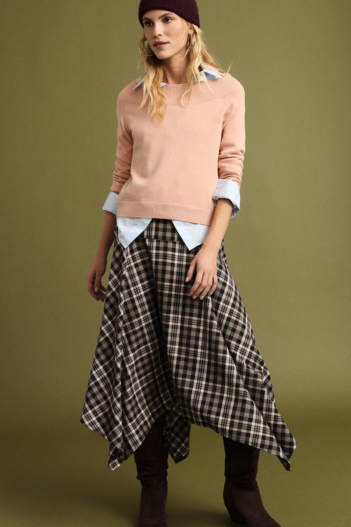 Female model in pink sweater, blue shirt and asymmetric black-and-white plaid skirt with brown boots and beanie on olive background