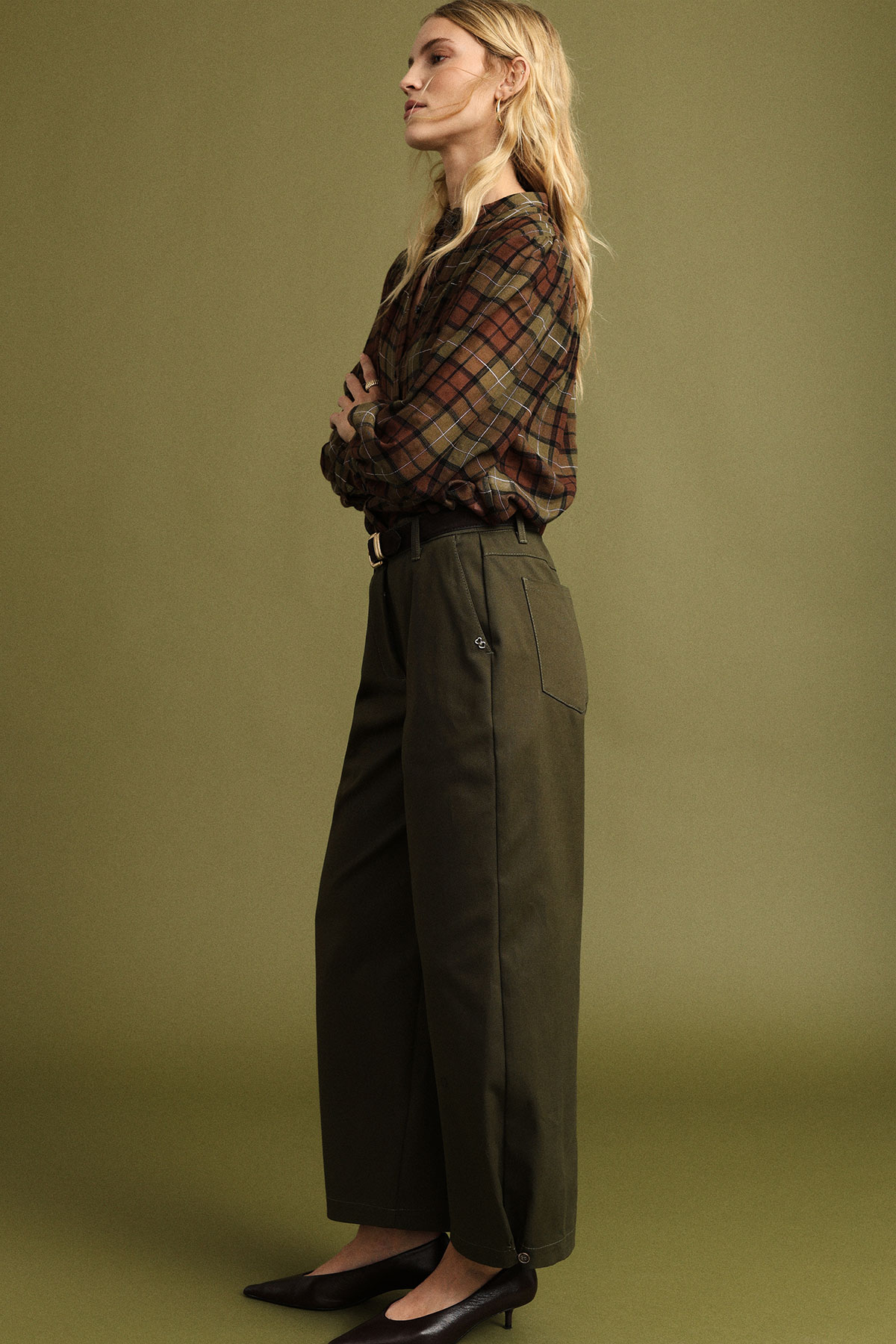 Side profile of woman in a brown plaid shirt and olive wide-leg trousers, arms crossed, wearing black heels against a green backdrop