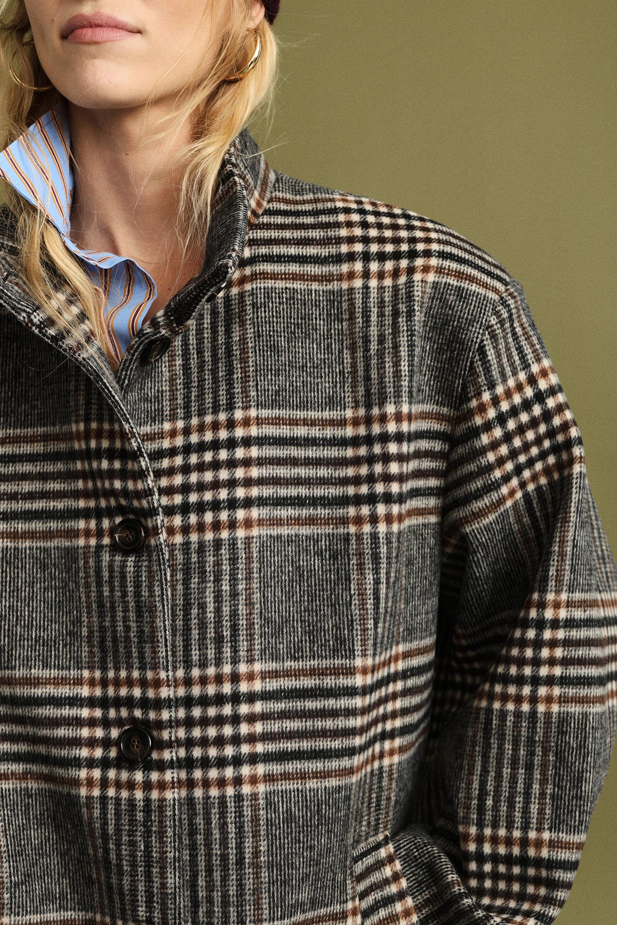 Cropped view of woman wearing grey and brown checked wool coat over blue striped shirt with gold hoop earring on an olive background