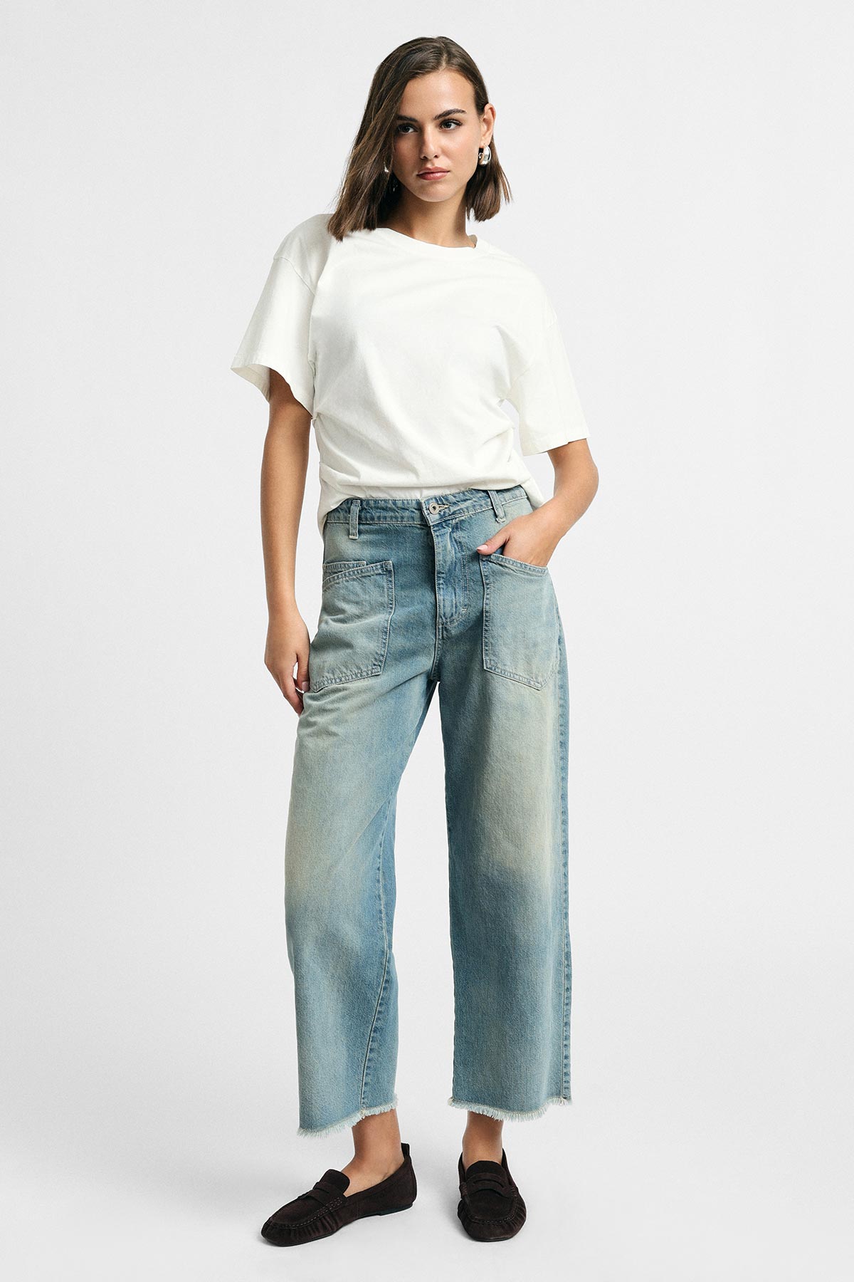 Model wearing white short-sleeve t-shirt and light-wash wide-leg cropped jeans with frayed hems, black loafers, hands in pockets