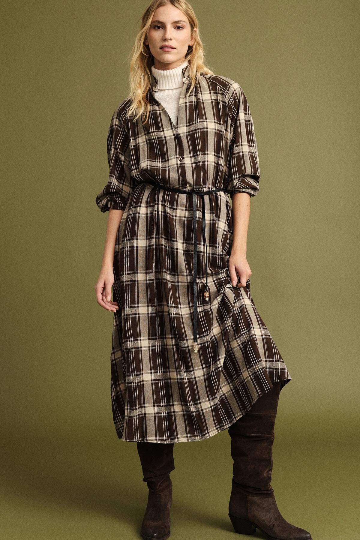 Woman in brown plaid belted midi shirt dress over cream roll-neck jumper, wearing knee-high brown suede boots against an olive background