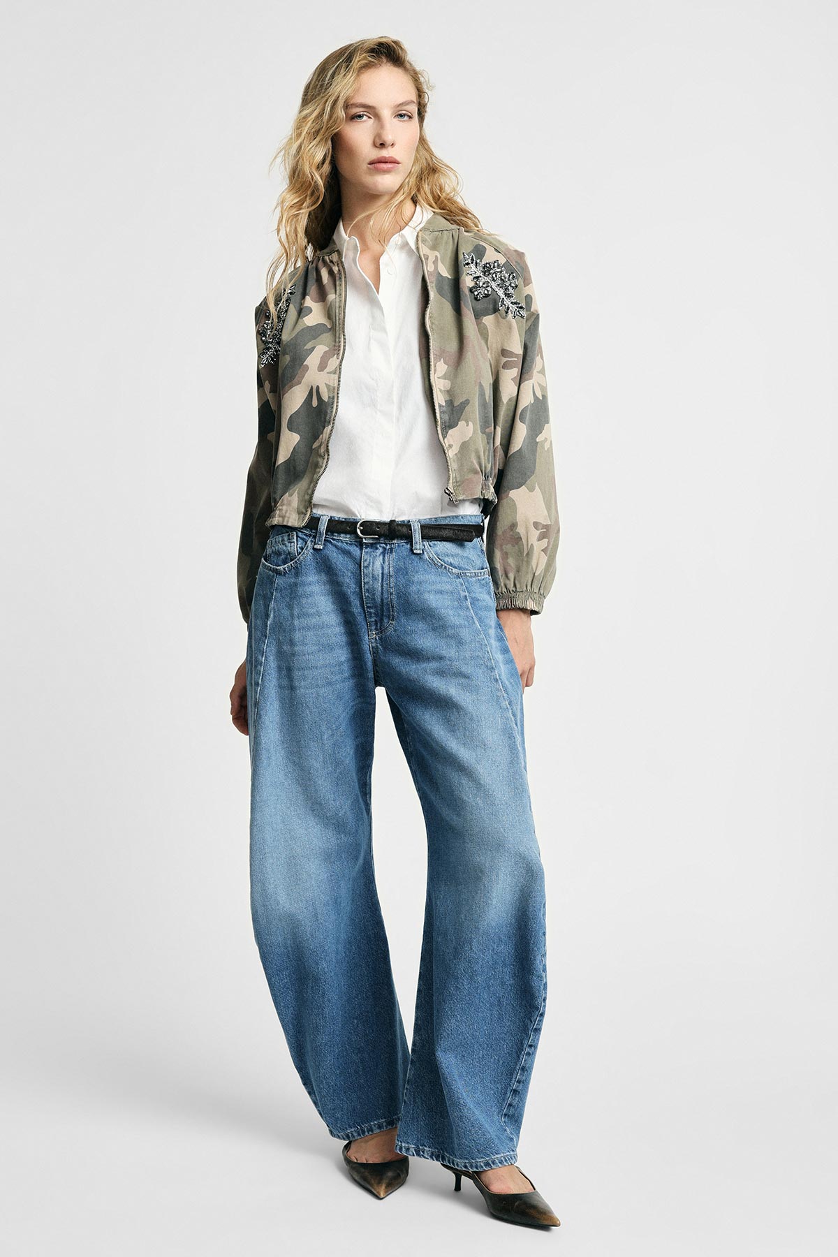 Woman in camouflage embroidered bomber jacket, white shirt, wide-leg blue jeans and black heels in full-length fashion pose