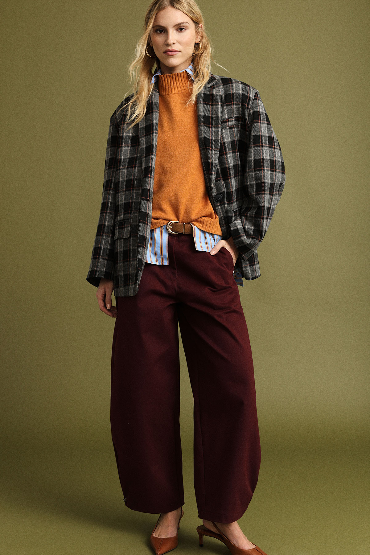 Model in checked blazer, orange knit and striped shirt with burgundy wide-leg trousers and brown heels on olive background