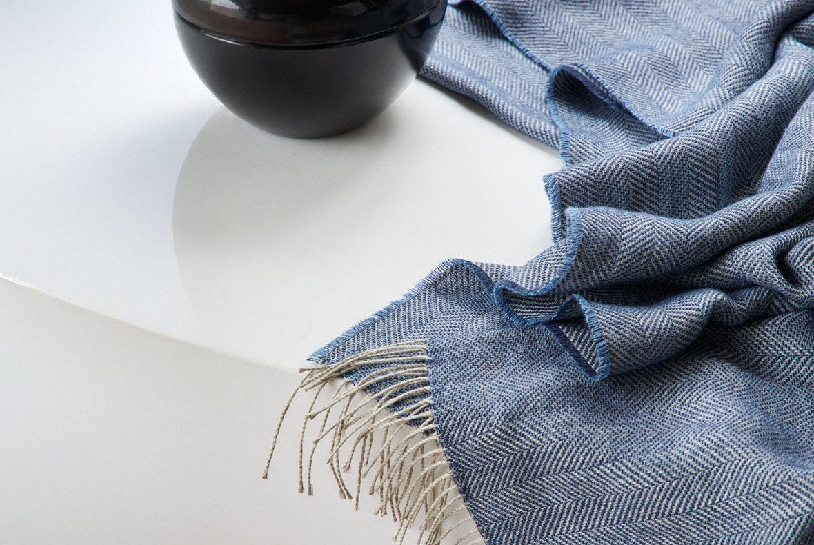 Blue herringbone woven throw with beige fringe draped on a white table beside a black ceramic bowl