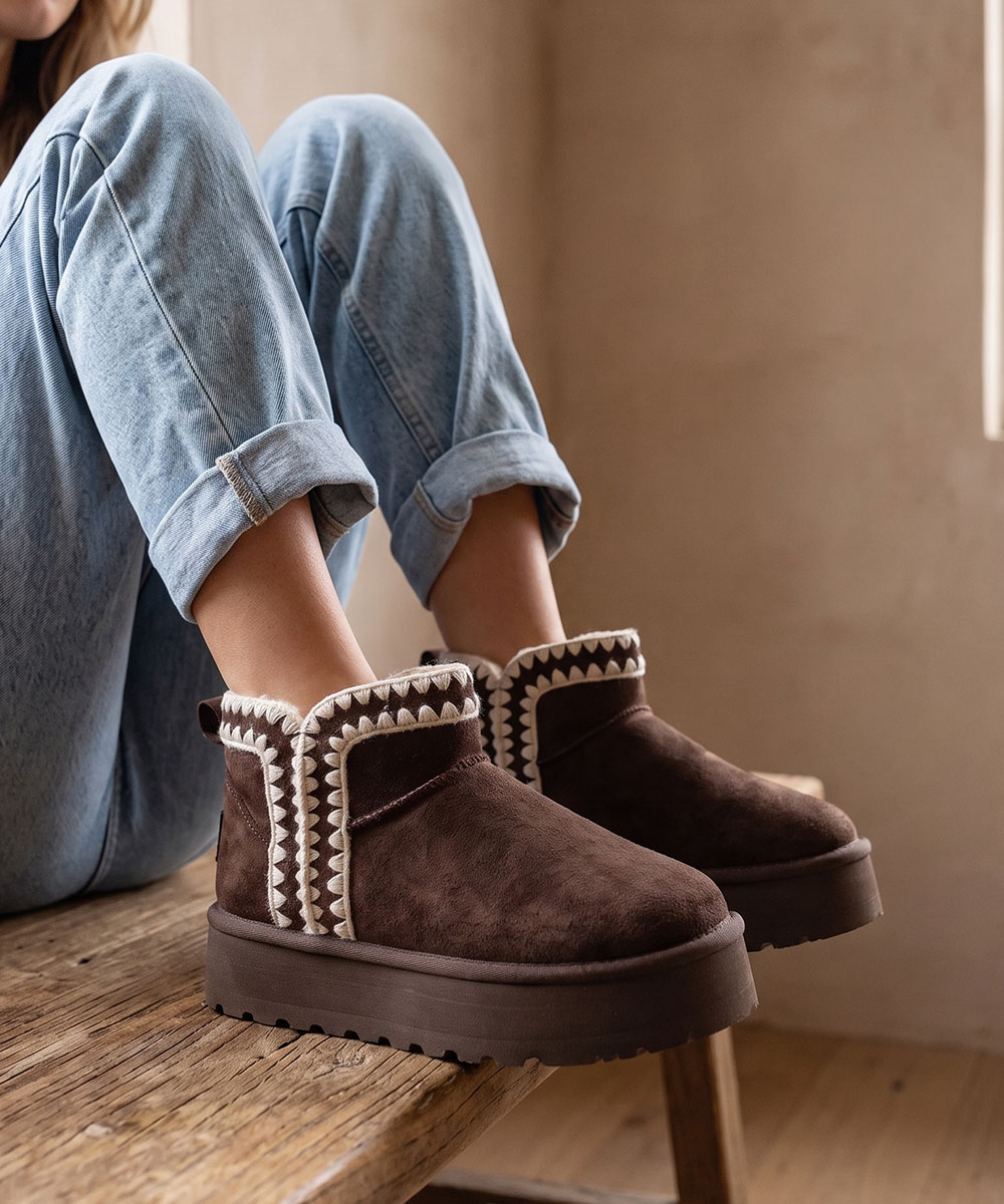 Brown suede platform ankle boots with white shearling trim worn with rolled-up light blue jeans on a wooden bench