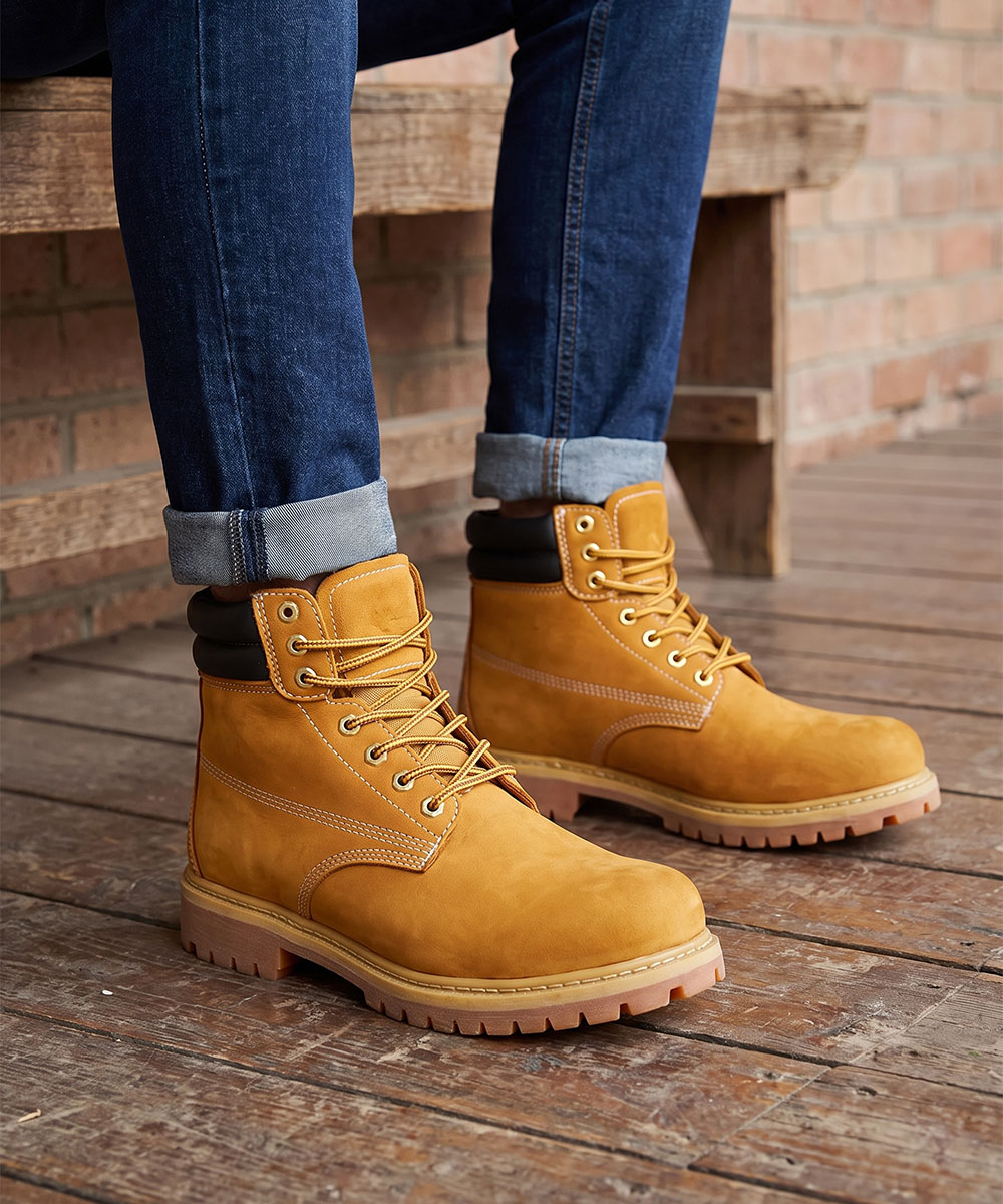 Tan lace-up work boots with padded collar and lug sole, worn with rolled blue jeans on wooden floor