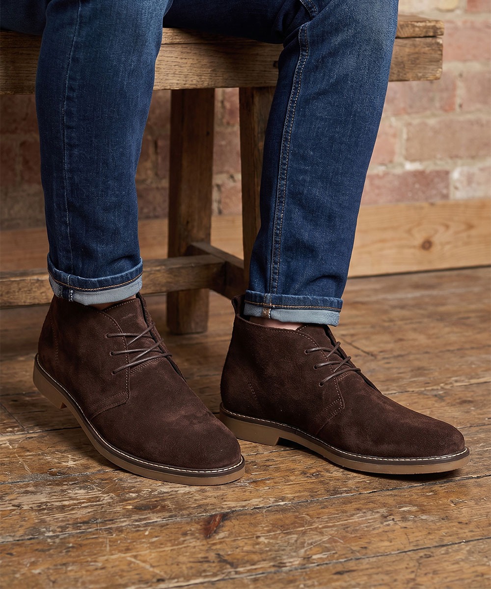 Men's brown suede chukka boots with cuffed blue jeans, person seated on a wooden bench over a rustic wooden floor