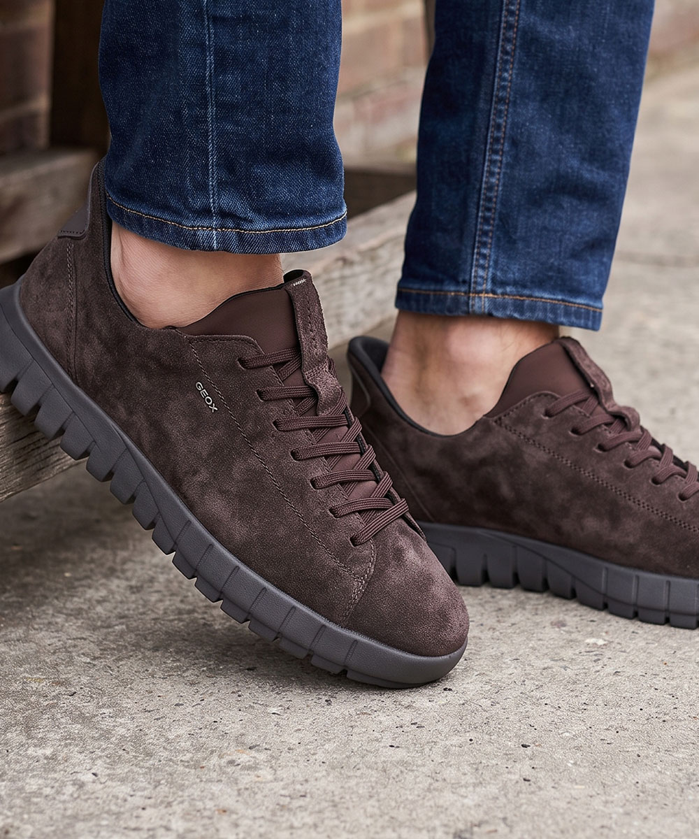 Men's brown suede trainers with chunky grey soles, worn with rolled blue jeans on a concrete step, casual urban footwear