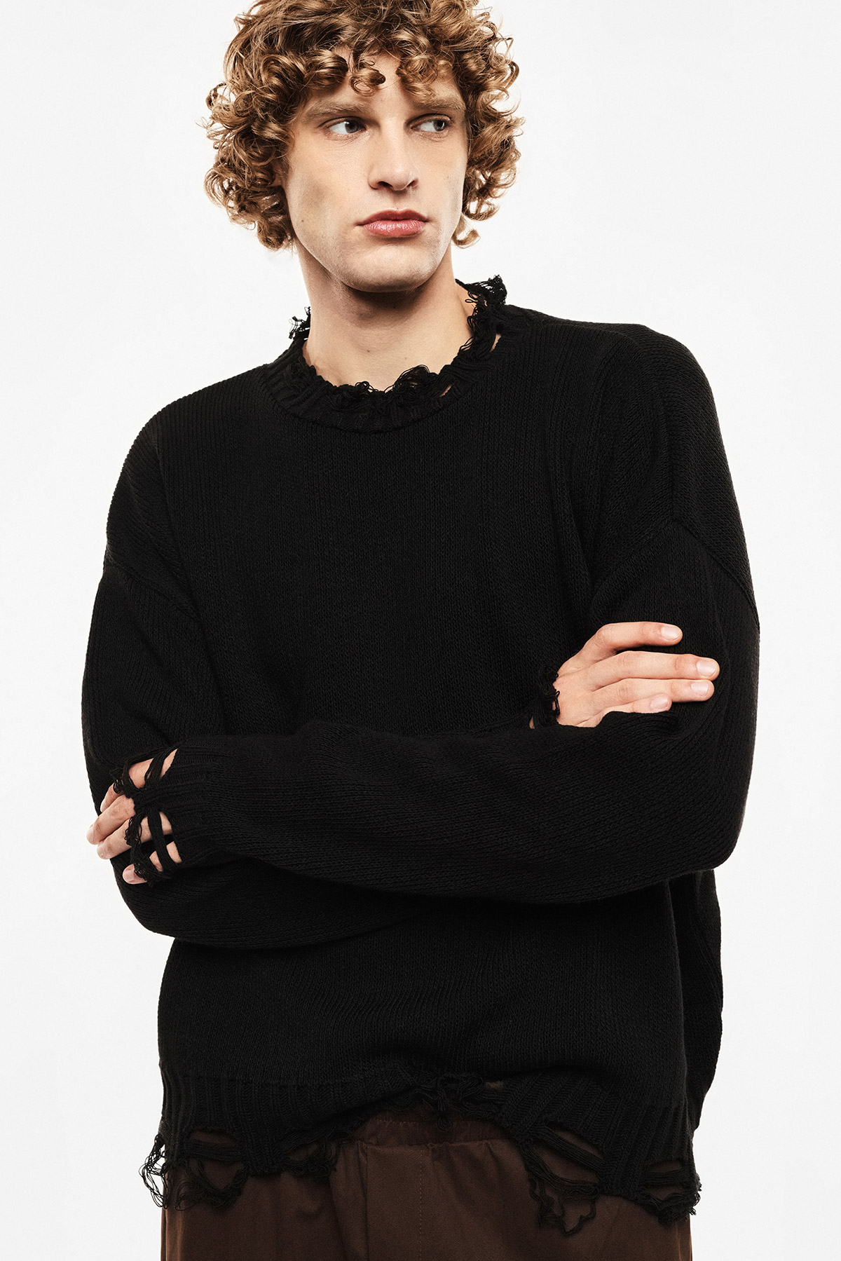 Young man with curly hair wearing a black distressed knitted jumper with frayed hems, arms crossed, looking to the side