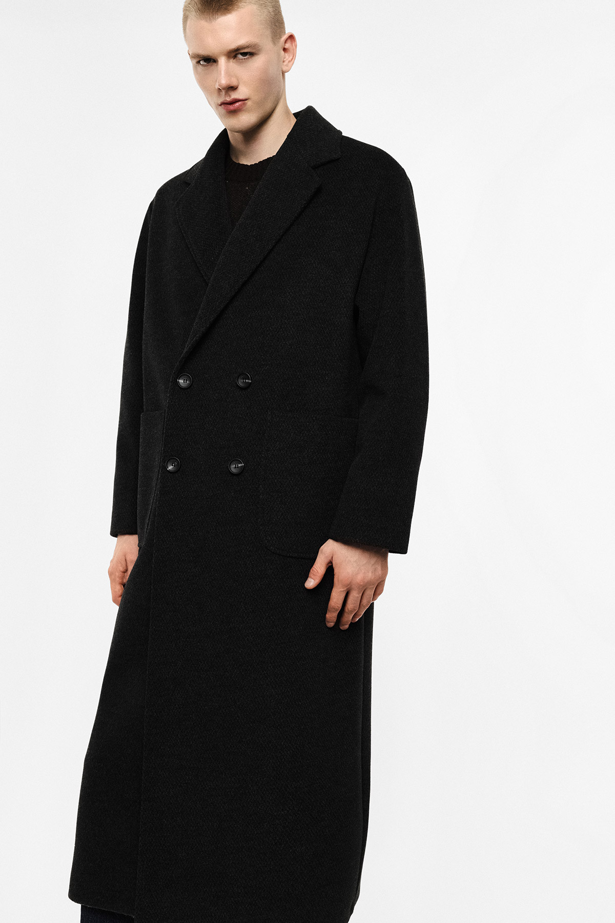Male model wearing long black double-breasted overcoat with notch lapel and patch pockets, standing against white background