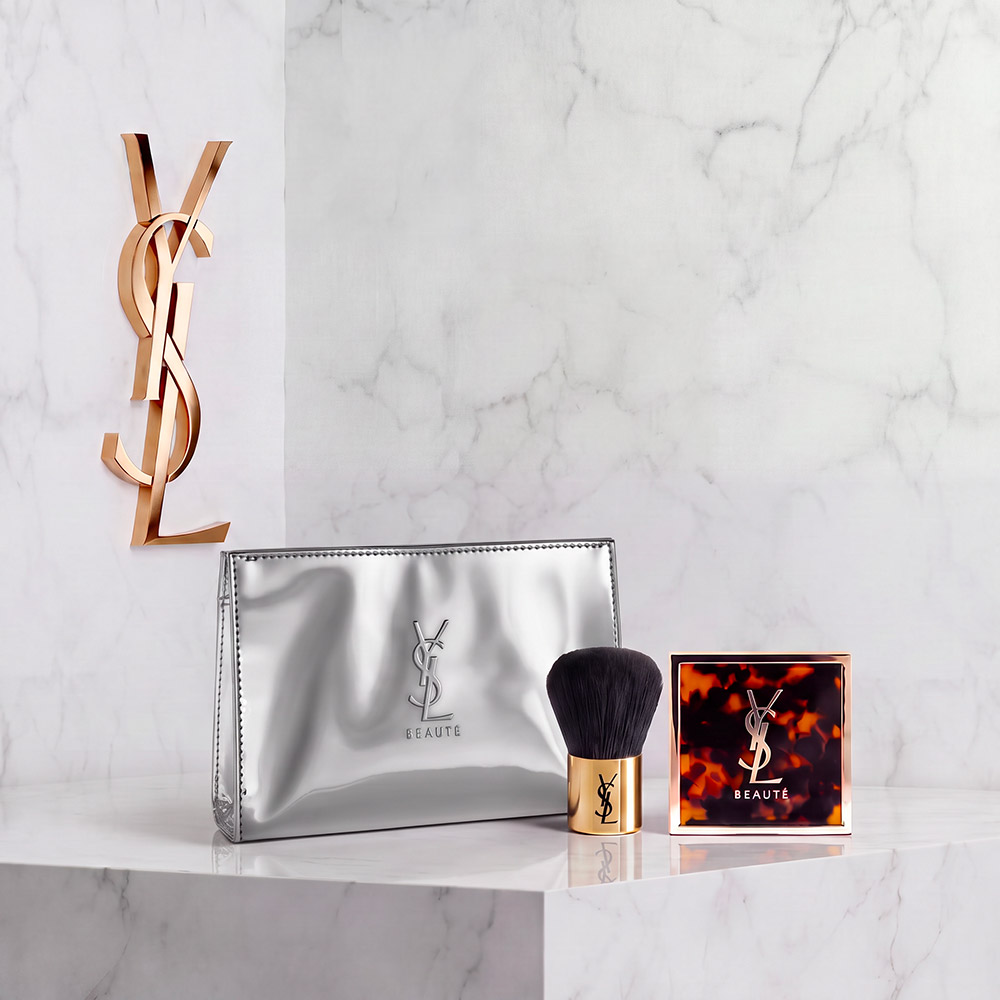 Set trucco YSL Beaut&eacute; con pochette argentata, pennello e compatto su uno sfondo effetto marmo.