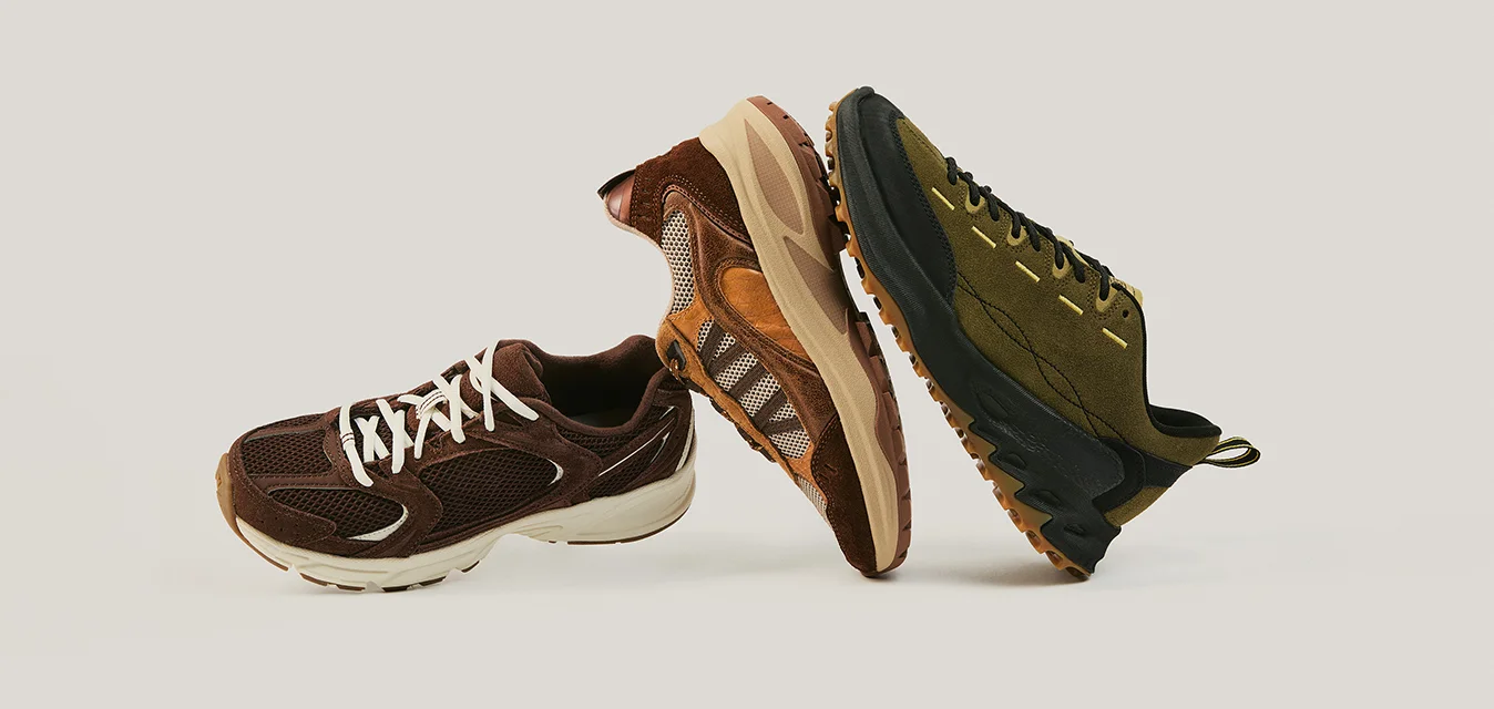 Tre scarpe da ginnastica nei colori marrone, sabbia e verde oliva disposte in piedi su uno sfondo neutro.