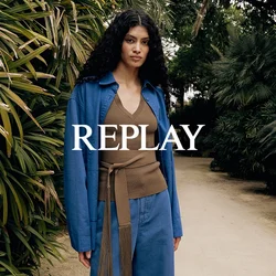 Donna con capelli ricci che indossa una giacca blu e pantaloni ampi, in piedi su un sentiero di terra circondato da piante tropicali dense e palme.