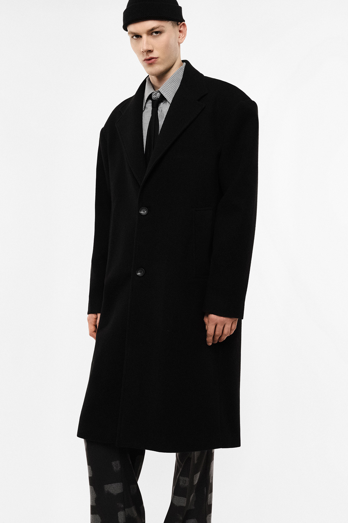 Man in long black overcoat with shirt, tie, patterned trousers and black beanie standing against white background