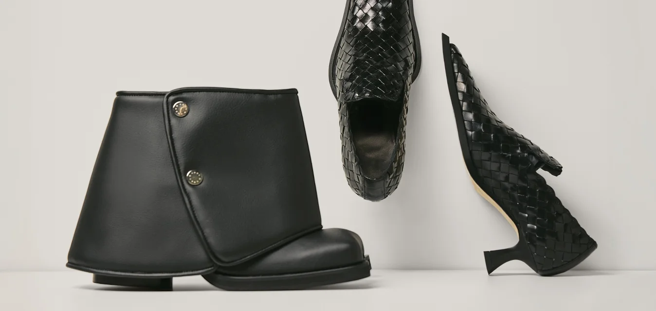 Due scarpe di pelle nera: un anfibio piatto con bottoni, un tacco alto a punta con texture intrecciata, mostrati su uno sfondo semplice.