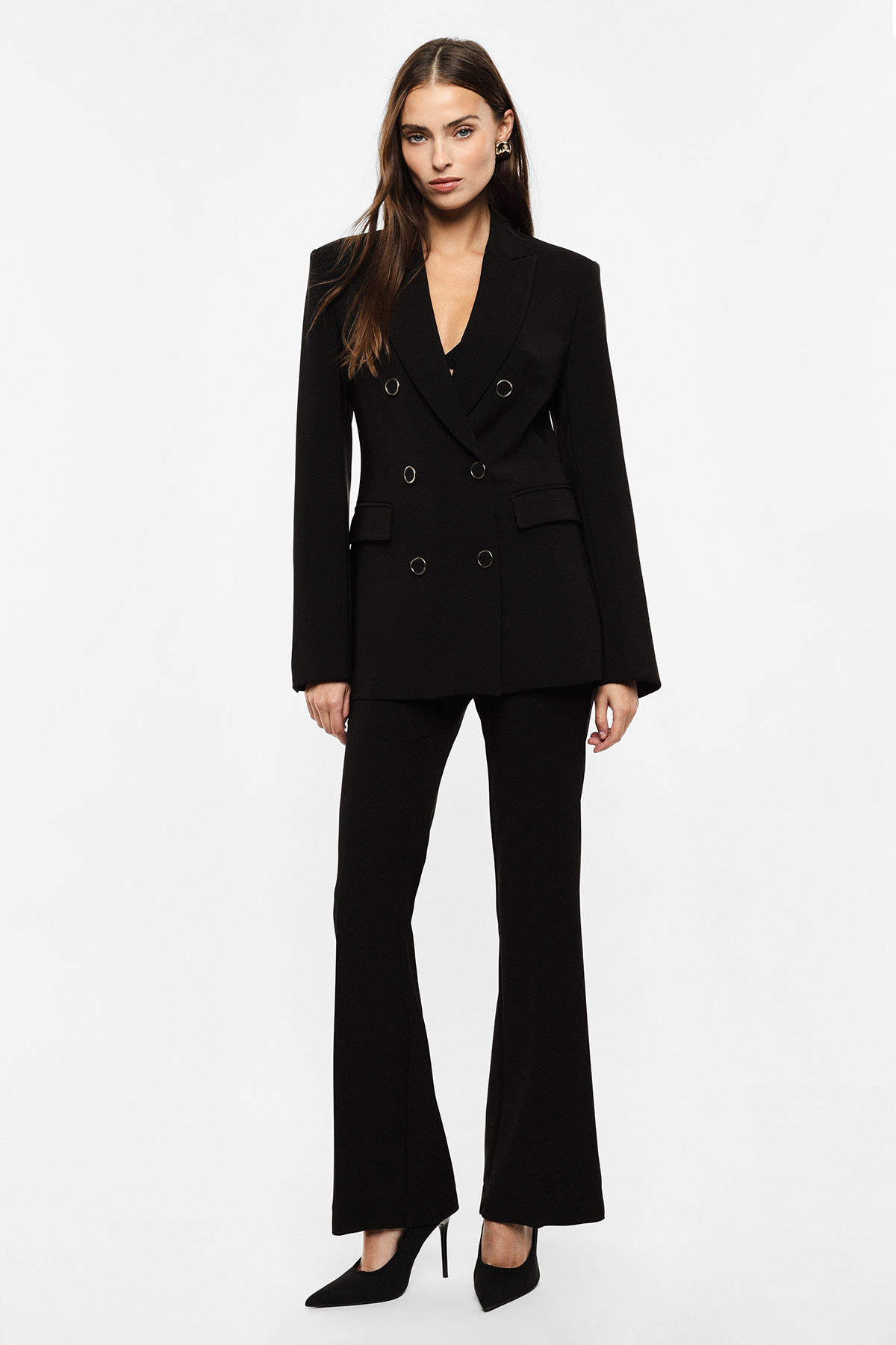 Female model wearing a tailored double-breasted black blazer and flared trousers with black heels, standing against a white background