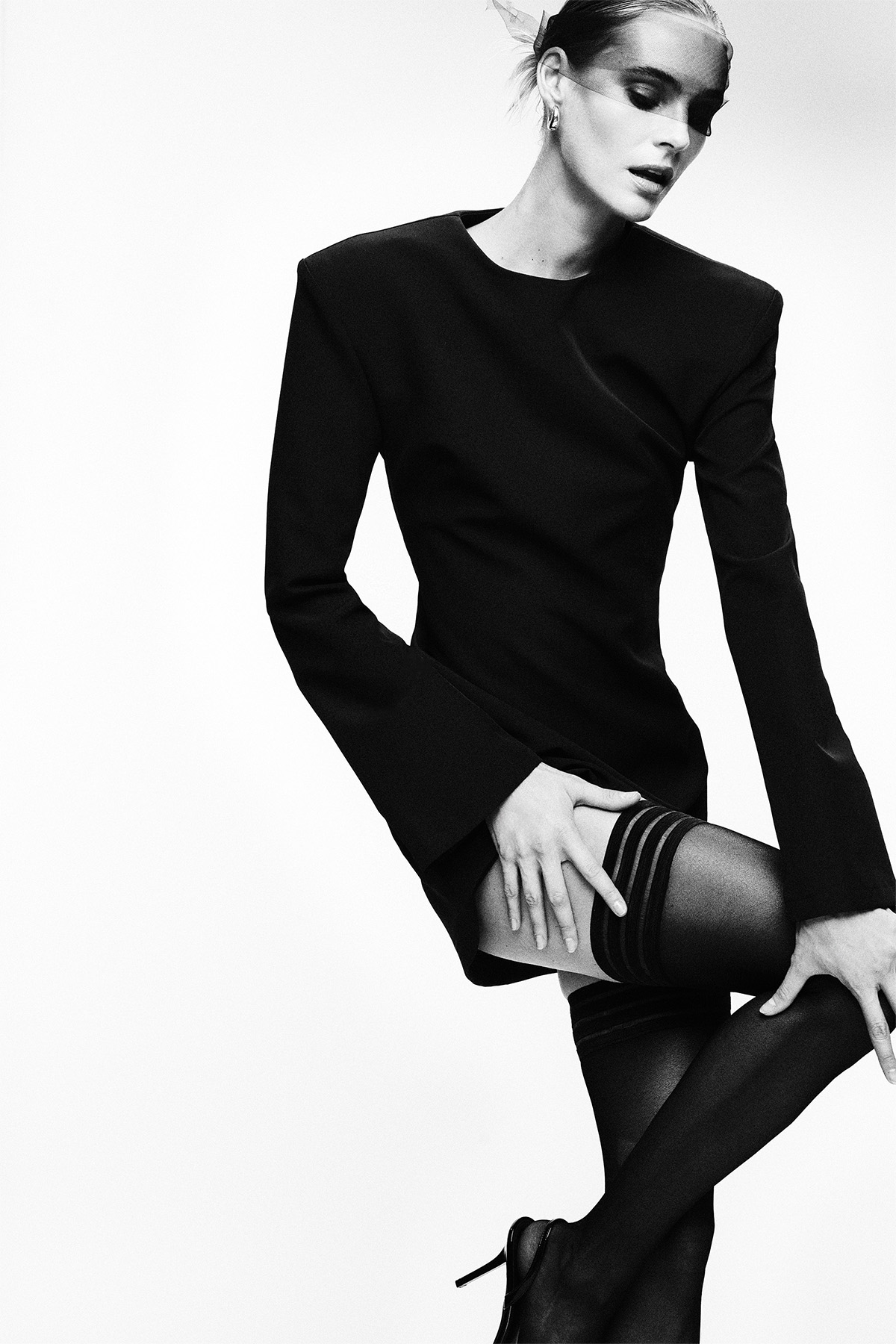 Black-and-white editorial of a model in a structured black blazer dress with thigh-high stockings and heels