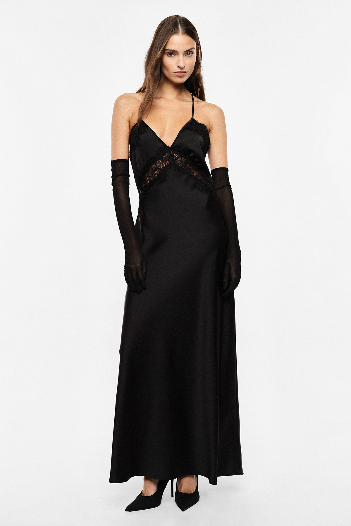 Woman wearing long black satin slip dress with lace trim, spaghetti straps, elbow-length sheer black gloves and black heels, standing