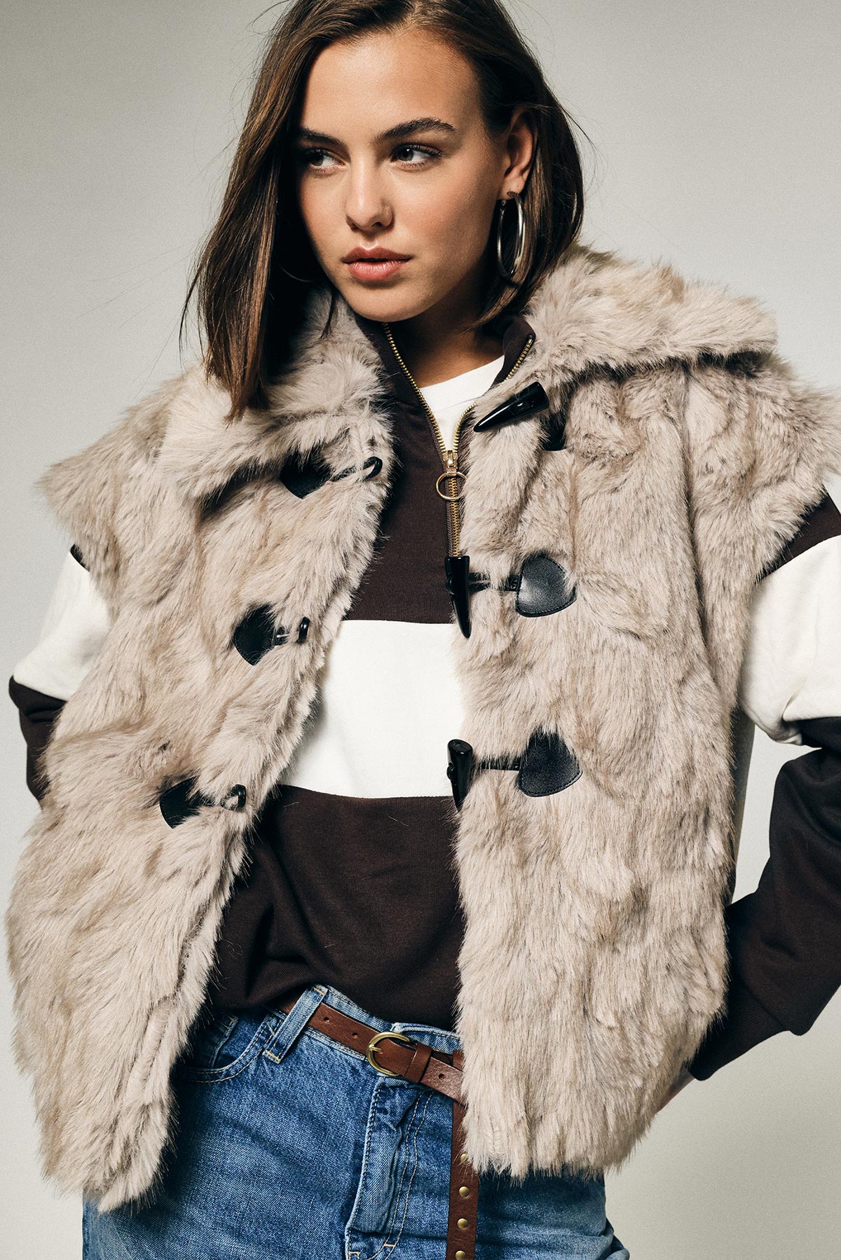 Young woman modelling beige faux fur sleeveless coat over striped jumper, hoop earring and jeans