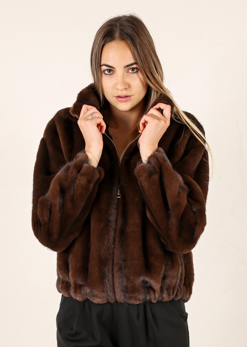 Model wearing a brown mink fur jacket, hands at the collar, posing against a plain light background for fashion photography