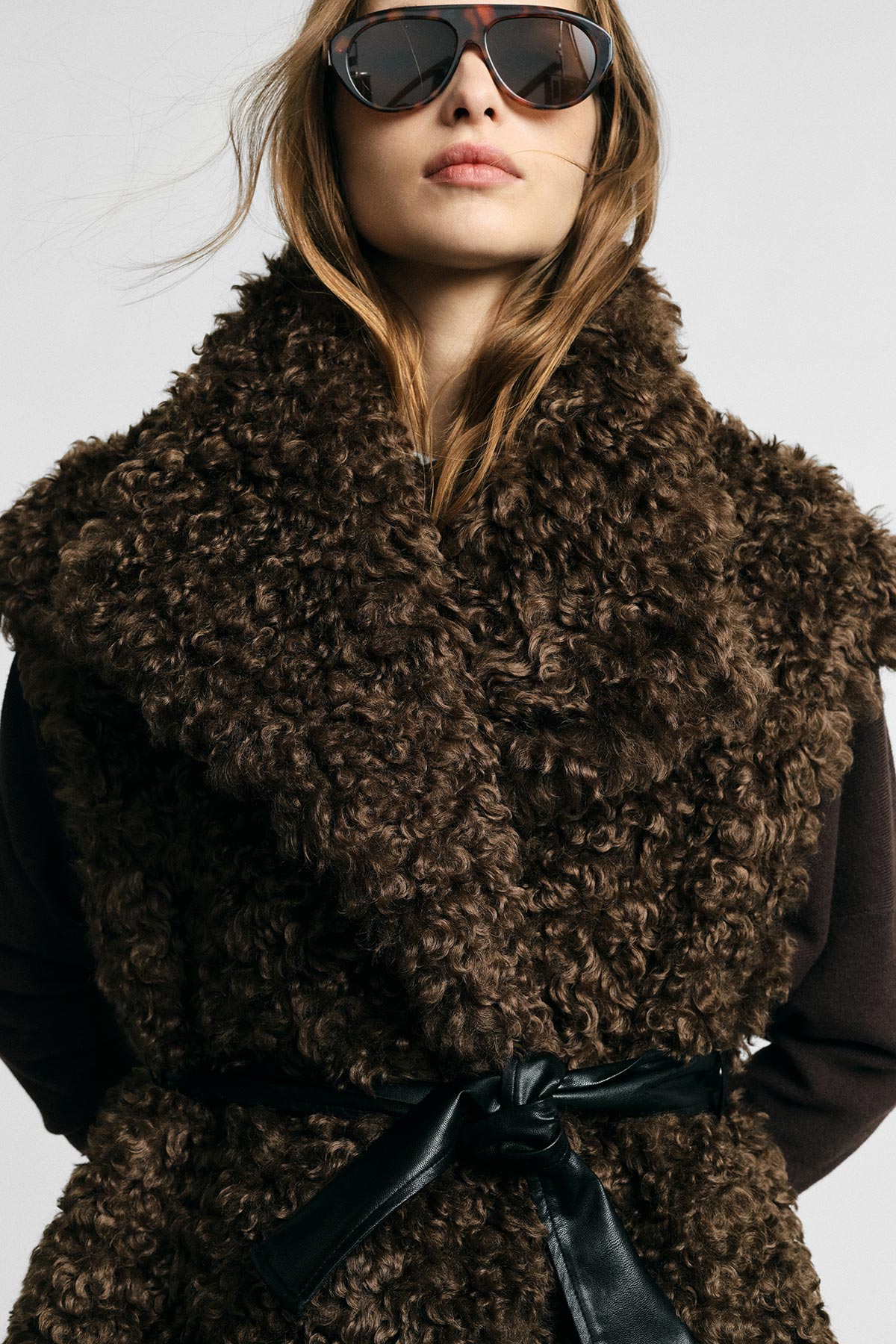 Close-up of model wearing dark brown curly shearling coat belted with black leather bow and sunglasses
