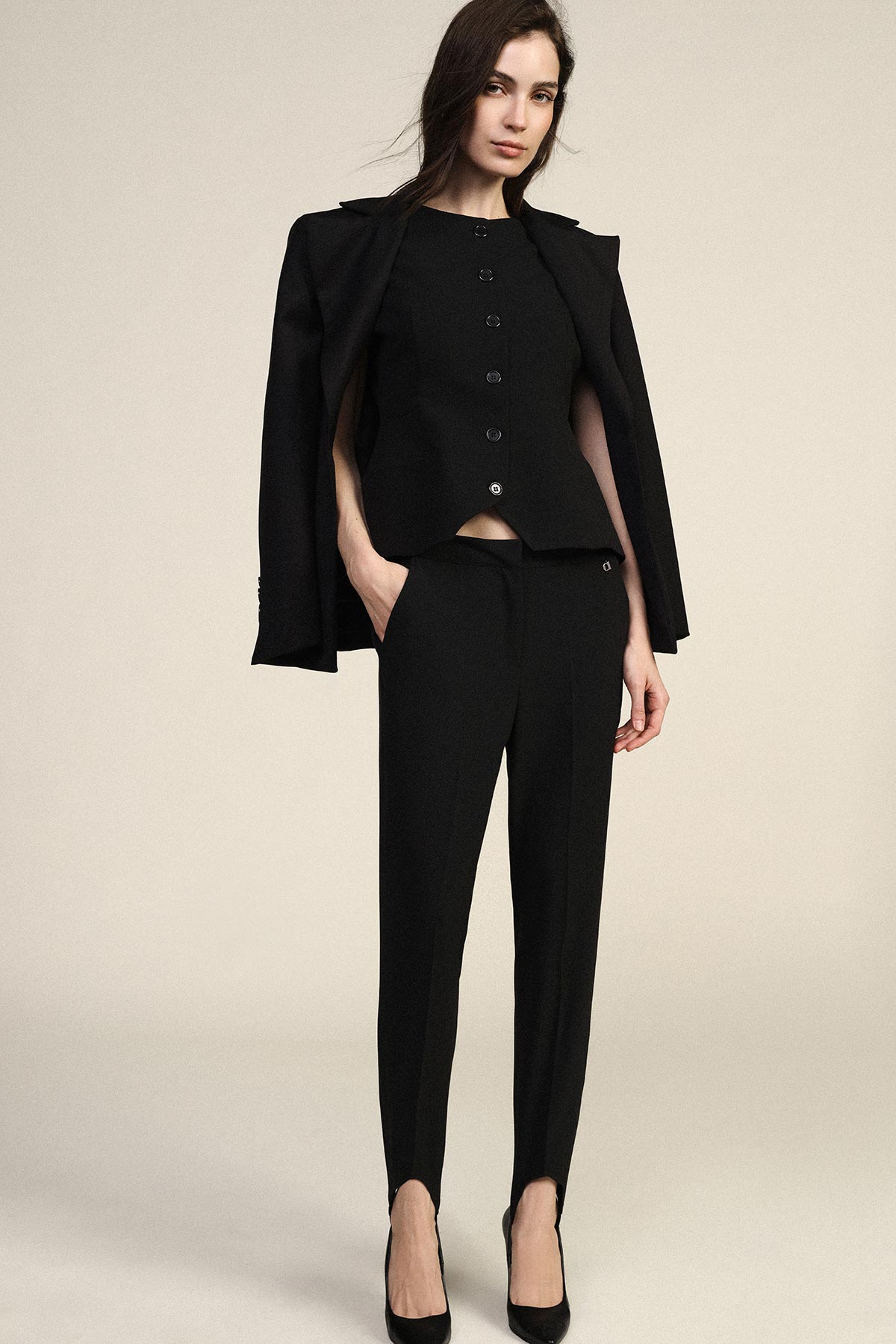 Woman in black tailored suit with jacket over shoulders, buttoned top, slim trousers and heels on neutral background