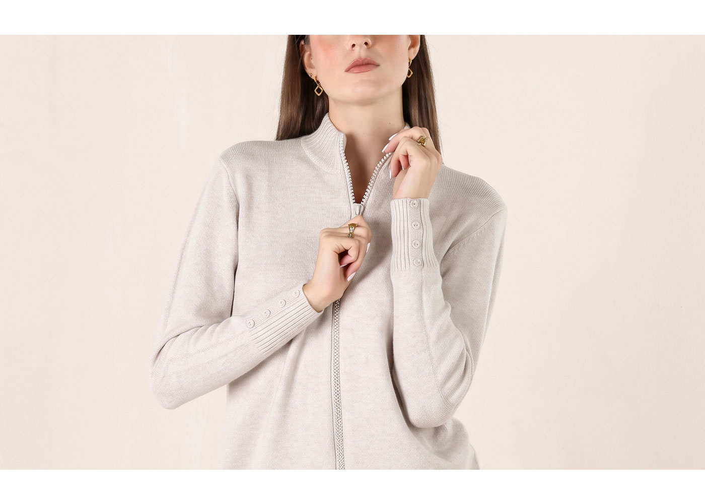 Woman adjusting beige zip‑up knitted cardigan with buttoned cuffs, wearing gold hoop earrings and rings
