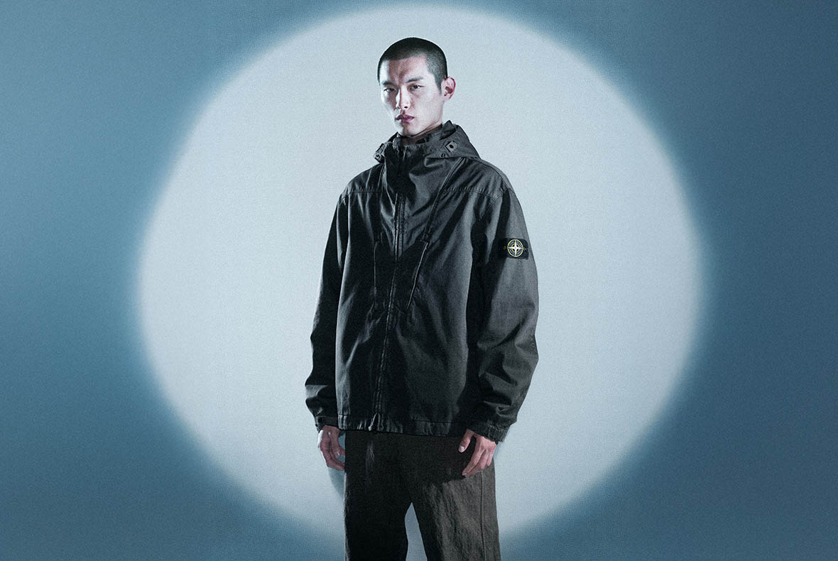 Young man standing in spotlight wearing dark hooded jacket with circular sleeve logo and brown trousers, neutral expression