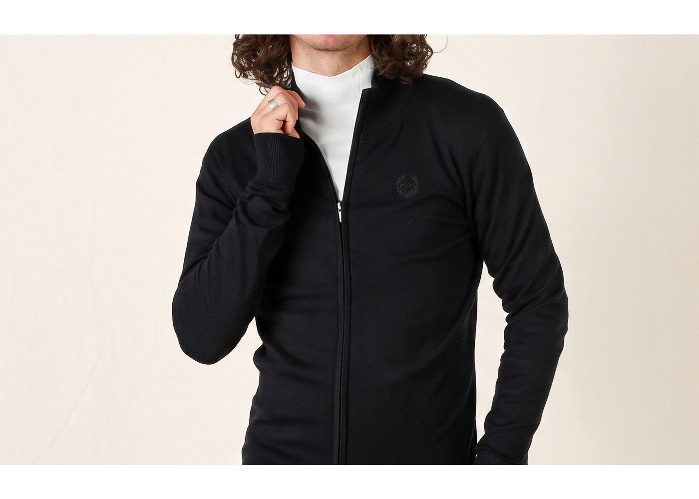 Person wearing black zip-up jacket over white roll-neck top, hand adjusting collar with cropped curly hair visible