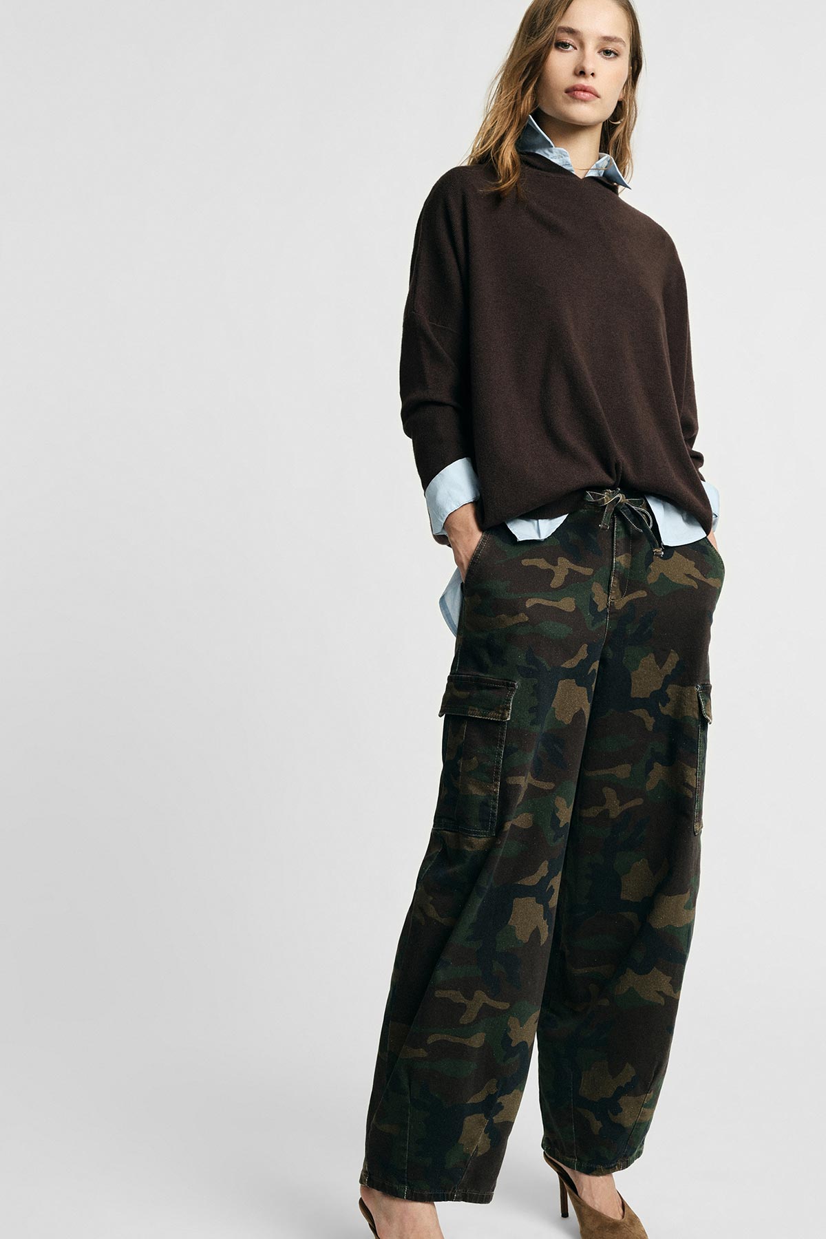 Woman standing against grey background wearing a brown oversized sweater over a blue shirt, camouflage cargo trousers and heels, hands in pockets