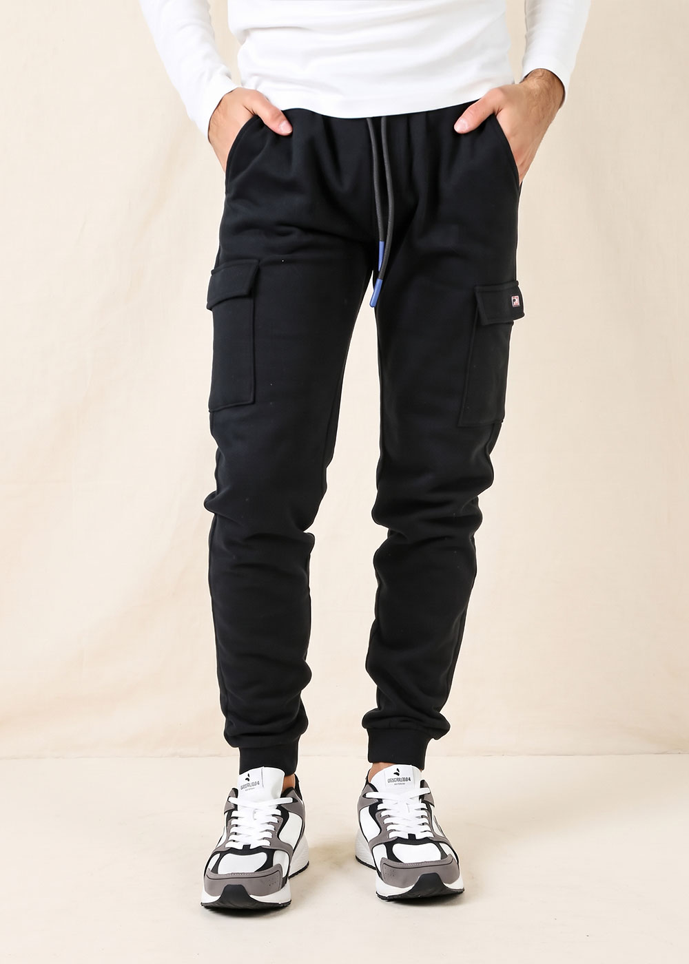 Model wearing black cargo joggers with drawstring waist, flap thigh pockets and cuffed ankles, paired with trainers.