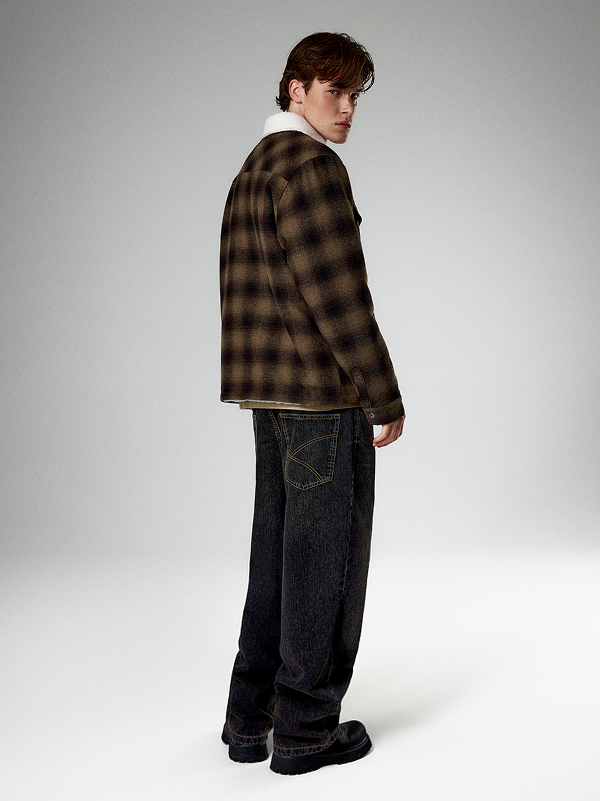 Uomo con giacca marrone a quadri, jeans scuri larghi e scarpe nere.