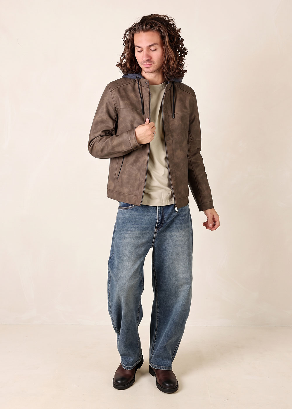 Young man wearing brown leather hooded jacket, beige T-shirt, loose blue jeans and brown boots, standing pose
