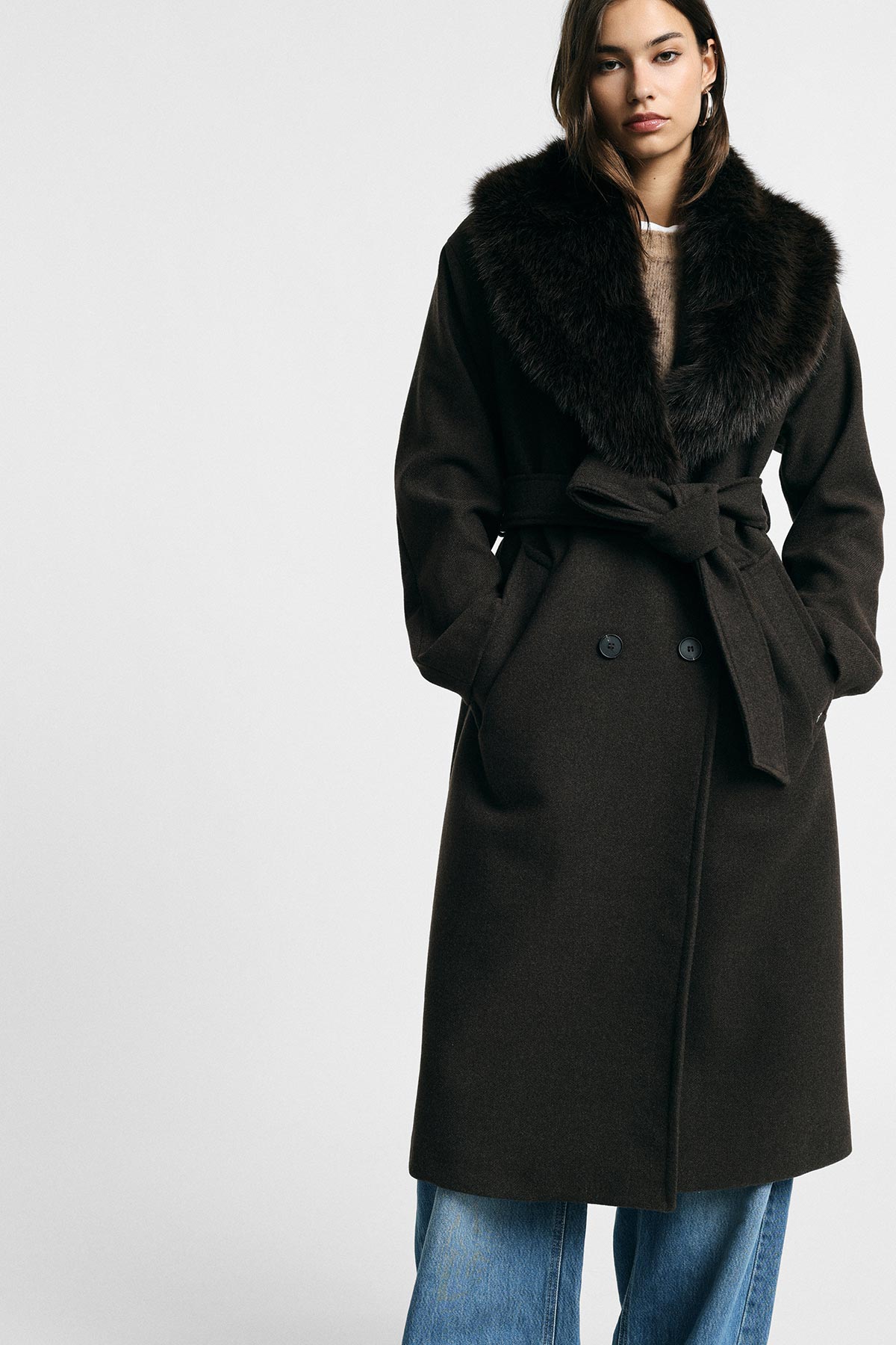 Woman modelling dark brown belted wool coat with large fur collar, hands in pockets, blue jeans, plain background