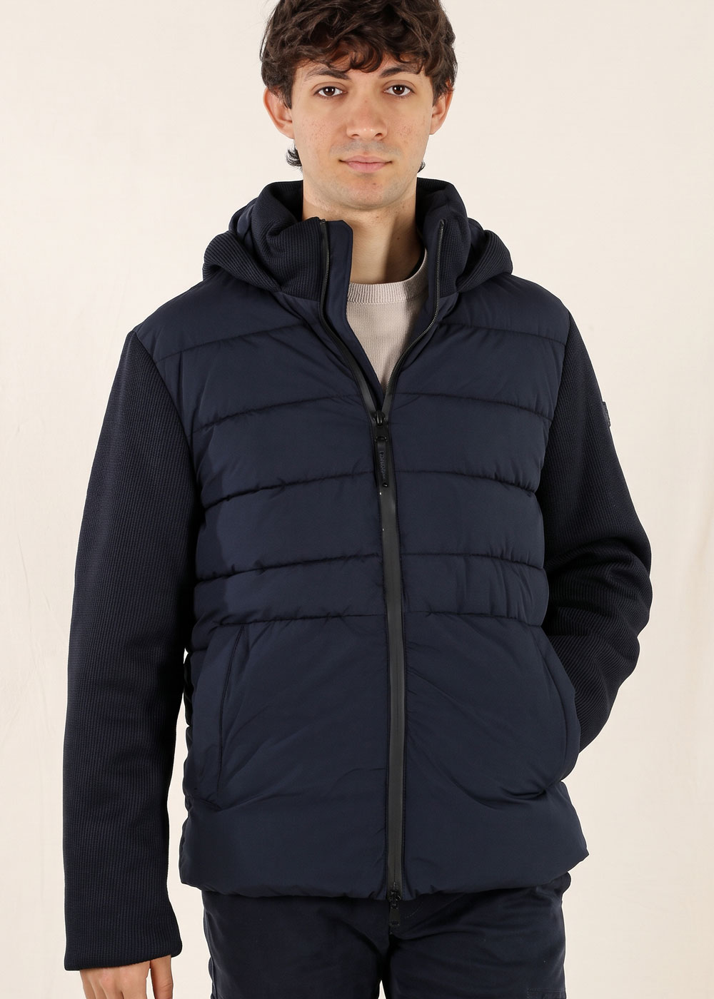 Man wearing a navy padded hooded jacket with ribbed knit sleeves and front zip, hands in pockets on a plain background