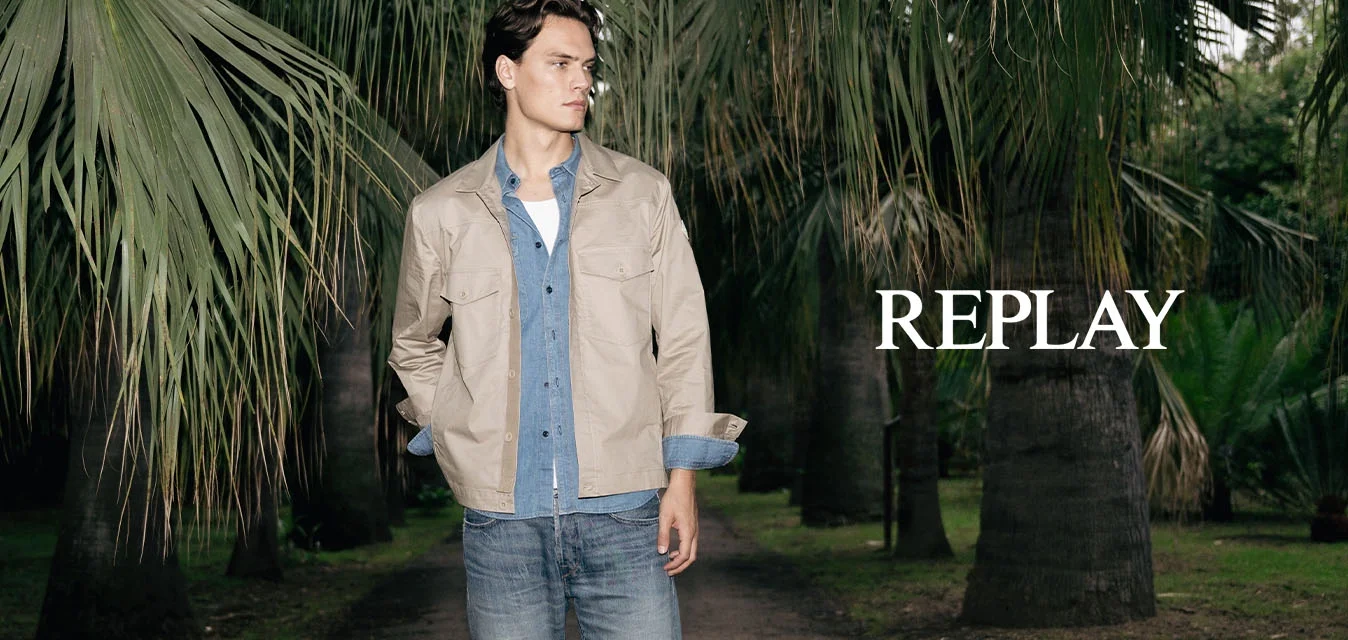 Giovane uomo in piedi su un sentiero sterrato in un'area boschiva, indossa una giacca beige, una camicia blu, jeans e scarpe grigie, guarda di lato.