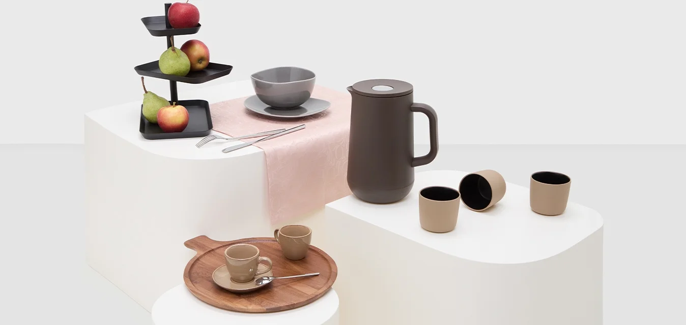 Ciotola grigia in ceramica, caraffa marrone scuro, vassoio in legno, tazze beige, porta frutta nera a strati con mele e pere su un runner da tavolo rosa.
