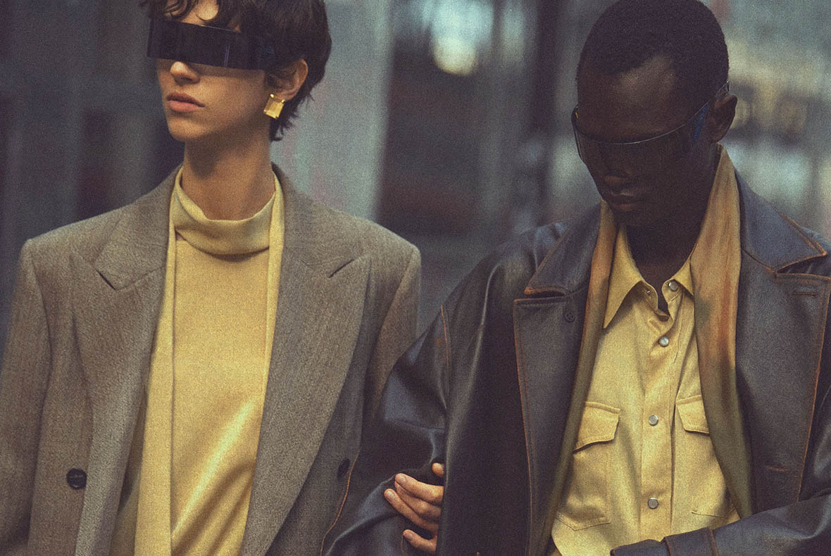 Two fashion models wearing oversized coats and futuristic sunglasses in muted tones, linked arms on an urban street