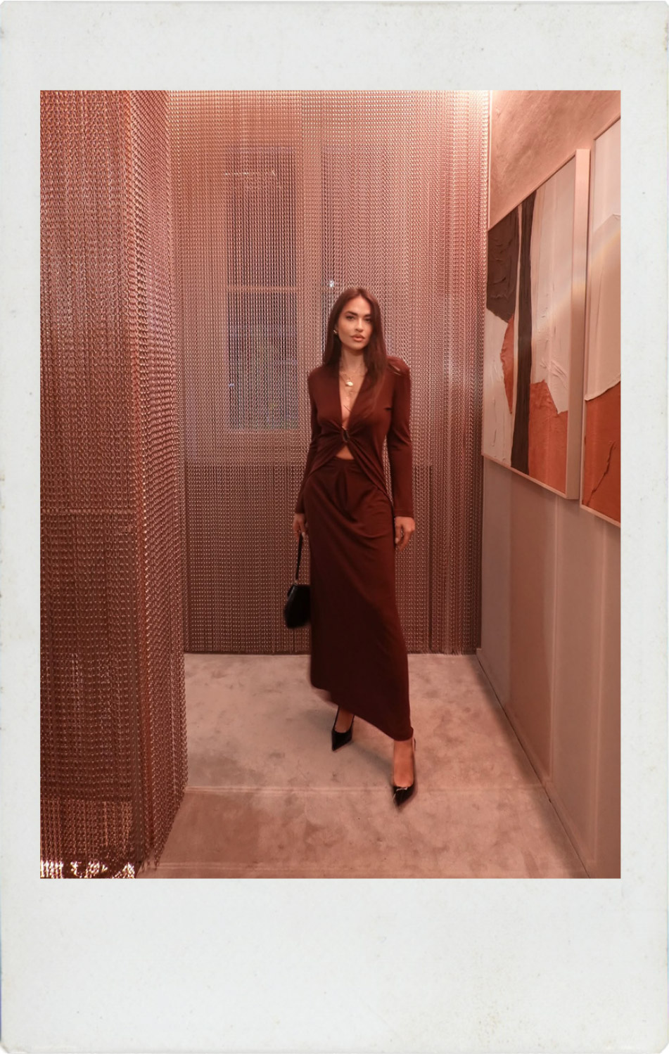 Woman in brown twist-front dress and black heels holding a small bag in a gallery corridor with metal chain curtain and abstract wall art