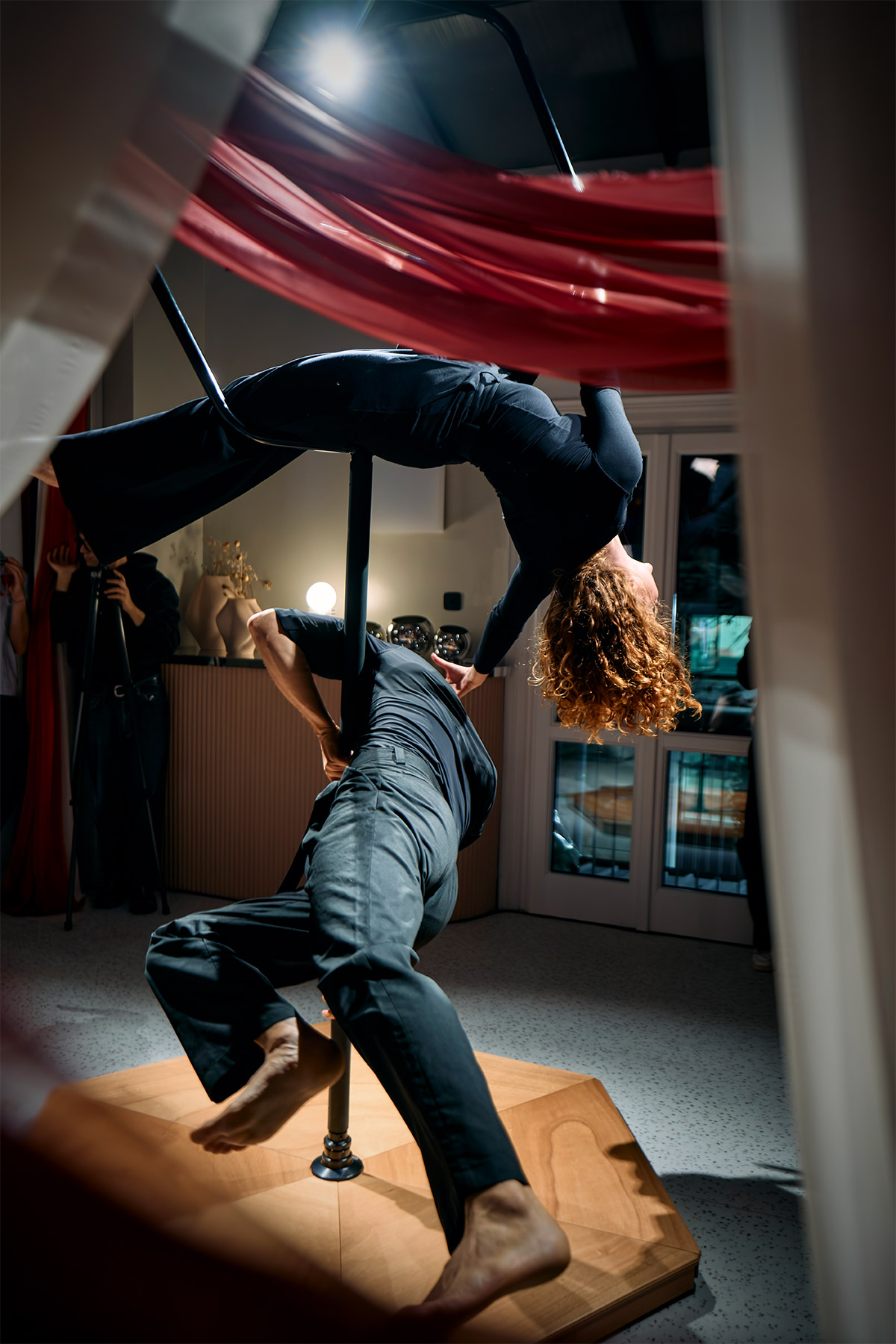 Two acrobats performing a pole and aerial silk routine in a studio — woman inverted on the pole beneath red fabric, male partner supporting