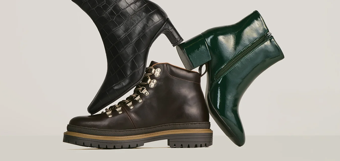 Tre stivali alla moda da donna in pelle con texture cocco nera, pelle marrone scuro con lacci e pelle verniciata lucida verde scuro, disposti in modo bilanciato su uno sfondo neutro.
