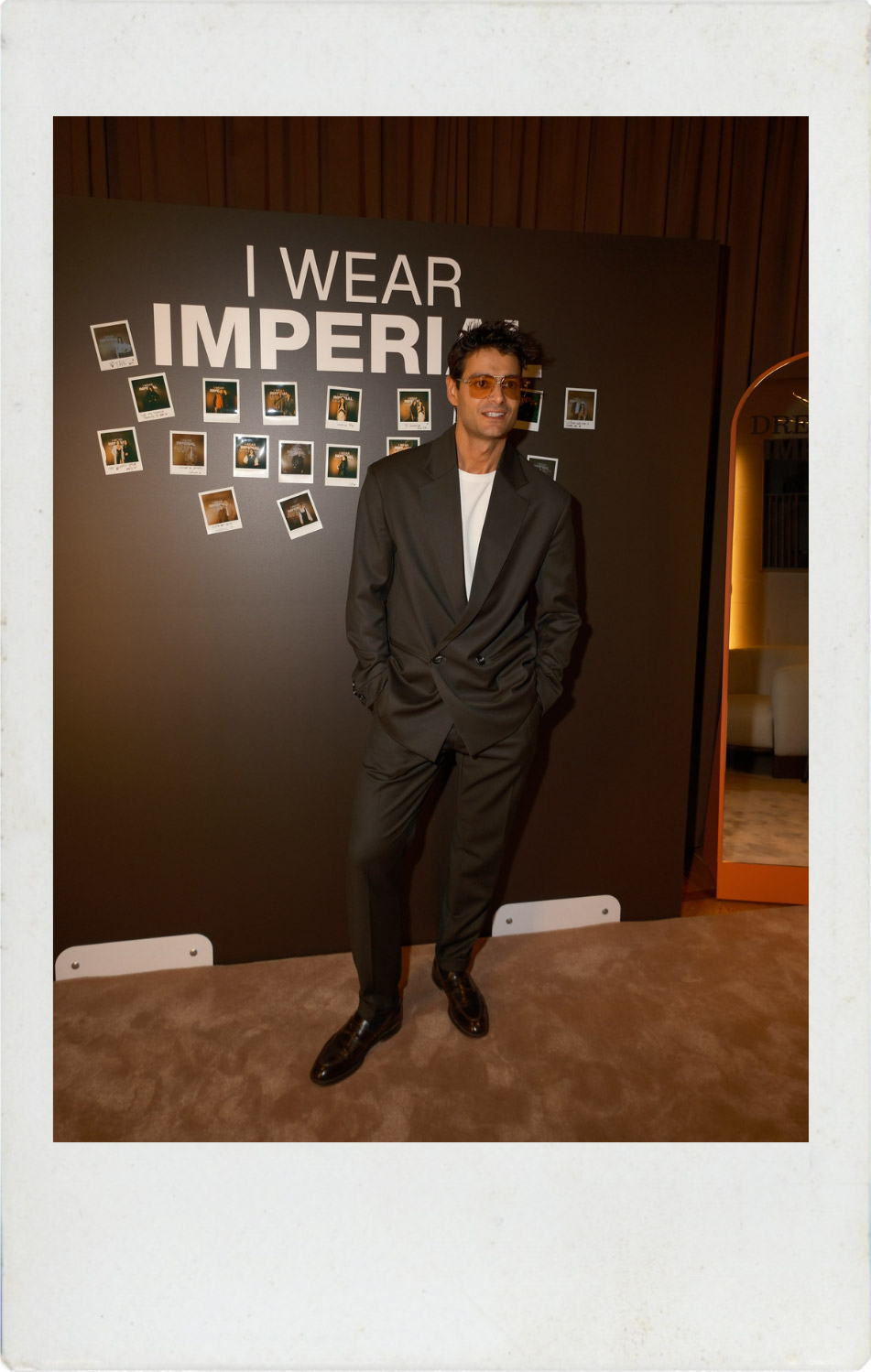 Man in dark suit and white T-shirt posing at a fashion event in front of an 'I WEAR IMPERI' wall decorated with instant photos