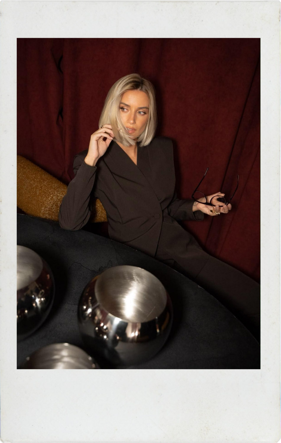 Blonde woman in dark tailored suit seated against red velvet curtain, holding spectacles, reflective silver spheres in foreground