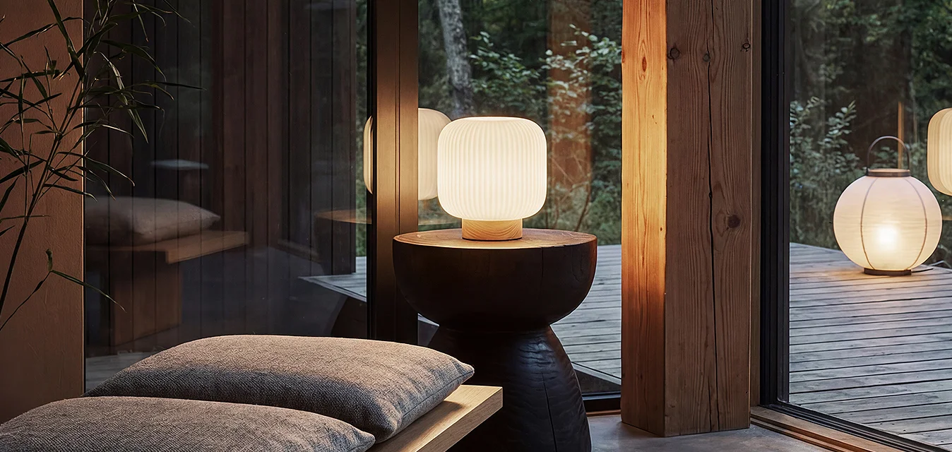 Area salotto interna moderna con cuscini, una lampada da tavolo laterale, grandi finestre di vetro che mostrano una lanterna esterna illuminata e gli alberi circostanti al crepuscolo.