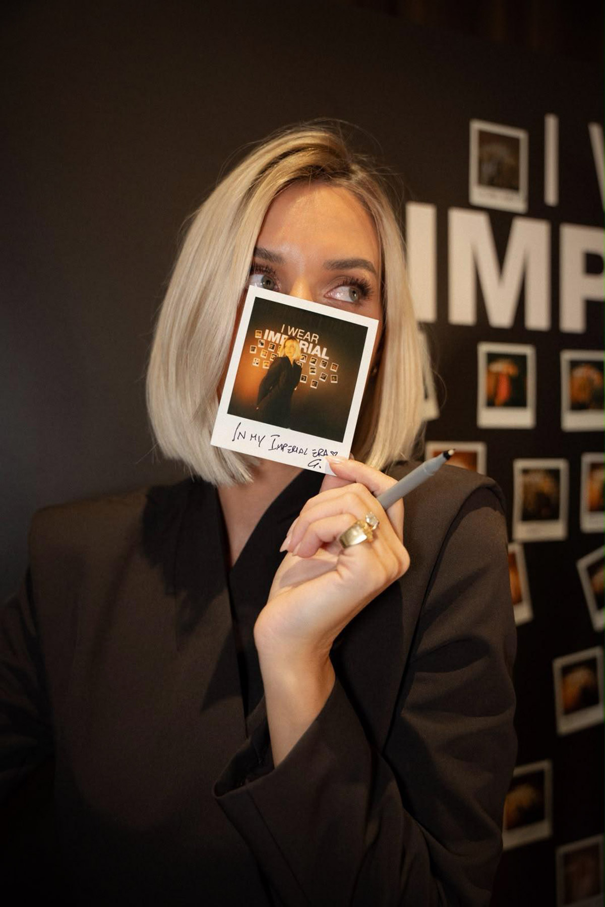 Blonde woman in a dark blazer holds a signed Polaroid over her mouth with a pen, photo wall backdrop