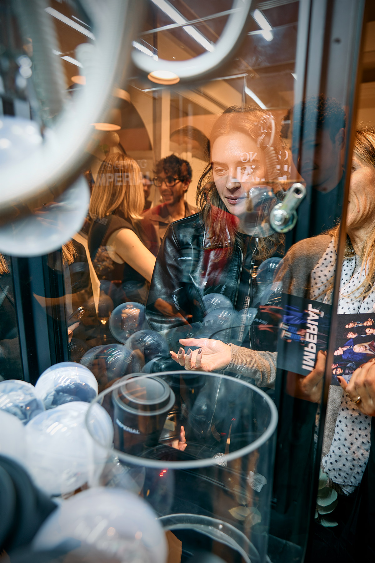 Young woman reaching into a glass prize machine to grab white plastic balls at a crowded indoor event, reflections visible