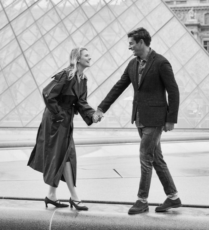 Smiling couple holding hands and walking in front of the glass pyramid entrance of the Louvre, woman in trench coat and heels, man in blazer